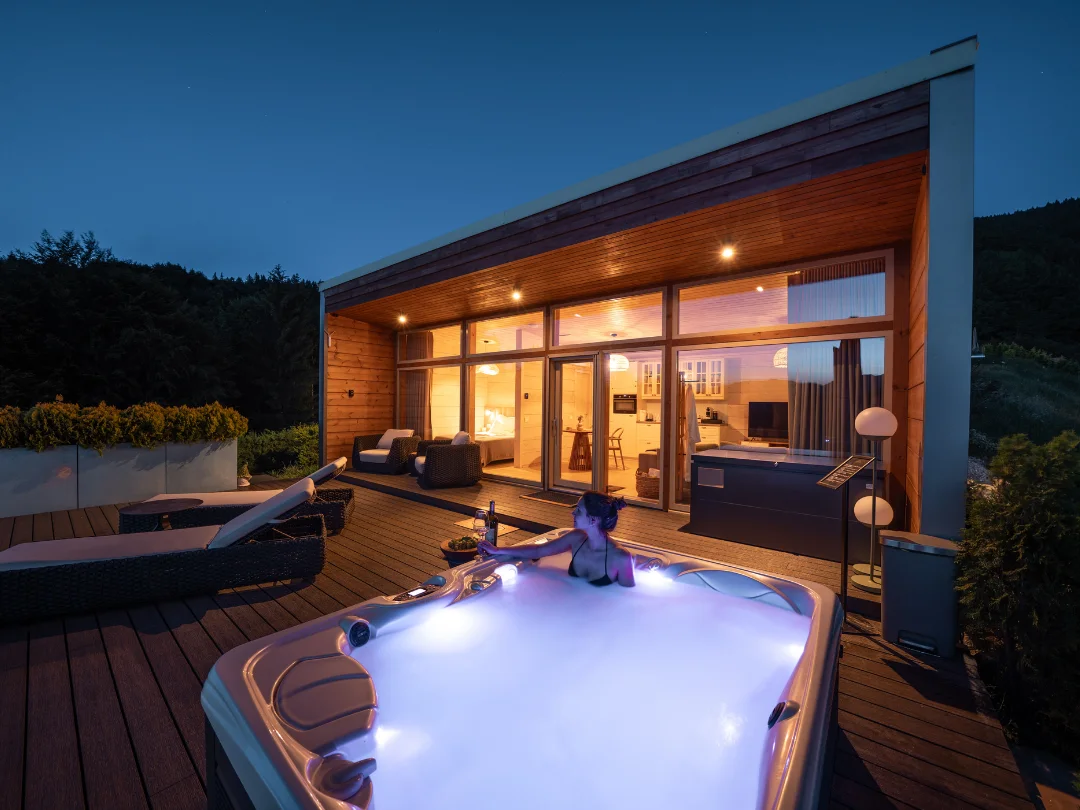 Wooden Eden Hill villa with illuminated windows and a woman in the terrace hot tub at night.