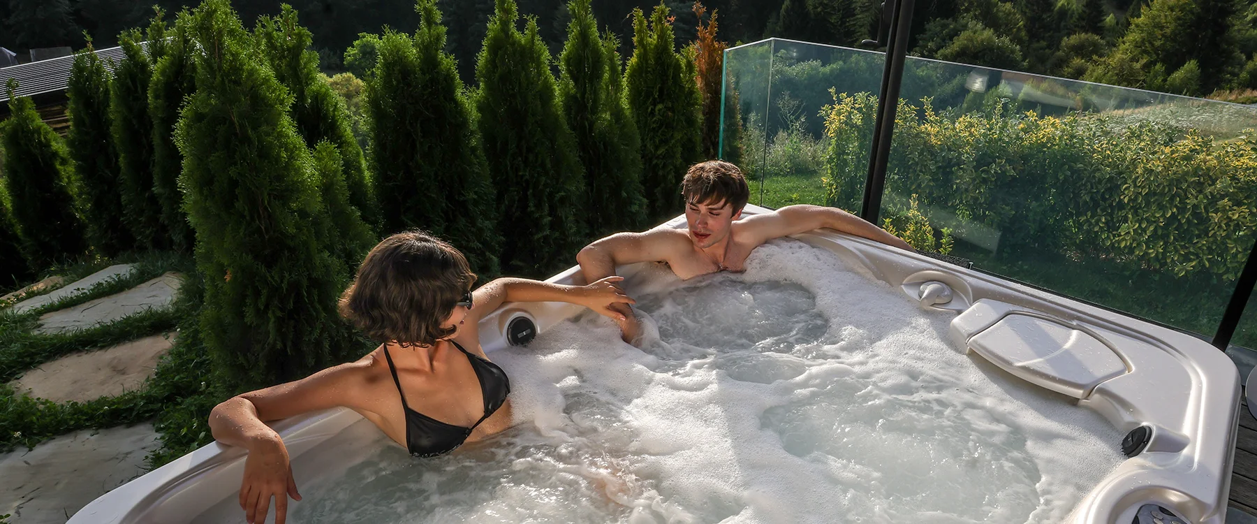 Couple facing each other in an Eden Hill terrace hot tub, with garden and forest behind them.