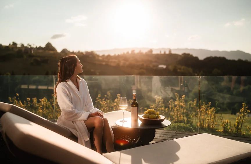 Woman in a robe on an Eden Hill terrace at sunset, beside a small table with a bottle, a glass and grapes.