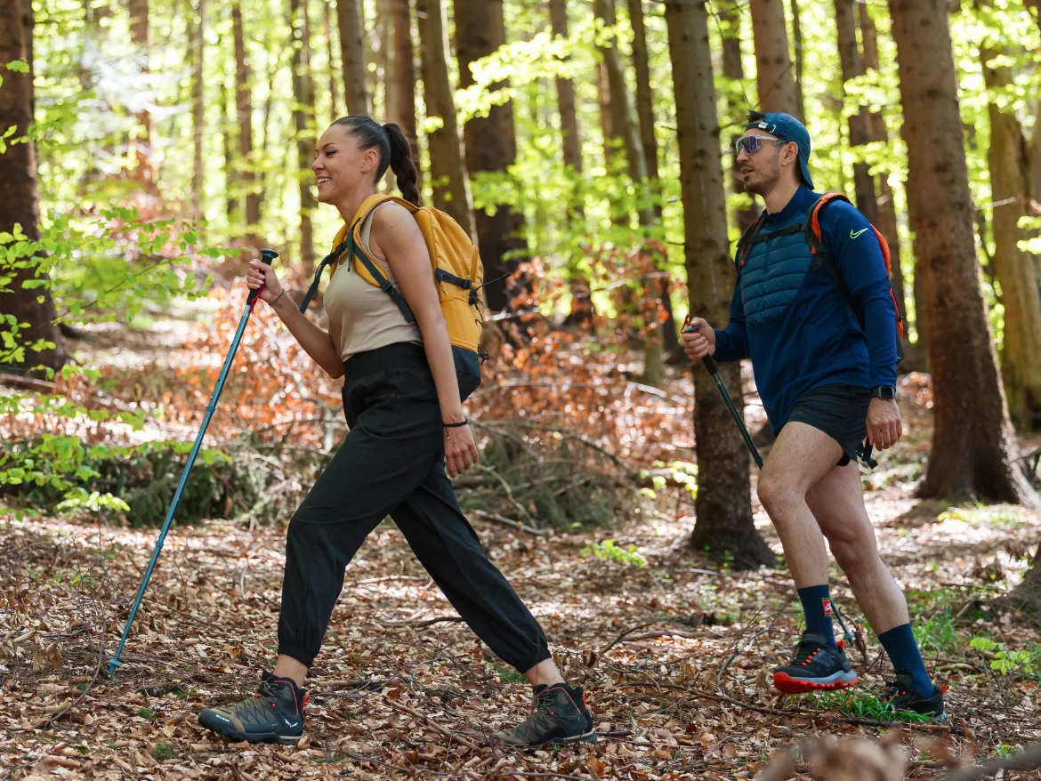 Two hikers with backpacks and trekking poles walking along a woodland path.