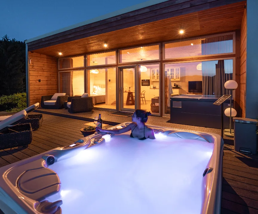 Femeie într-un jacuzzi iluminat în fața unei vile Eden Hill cu ferestre luminate, la lăsarea serii.
