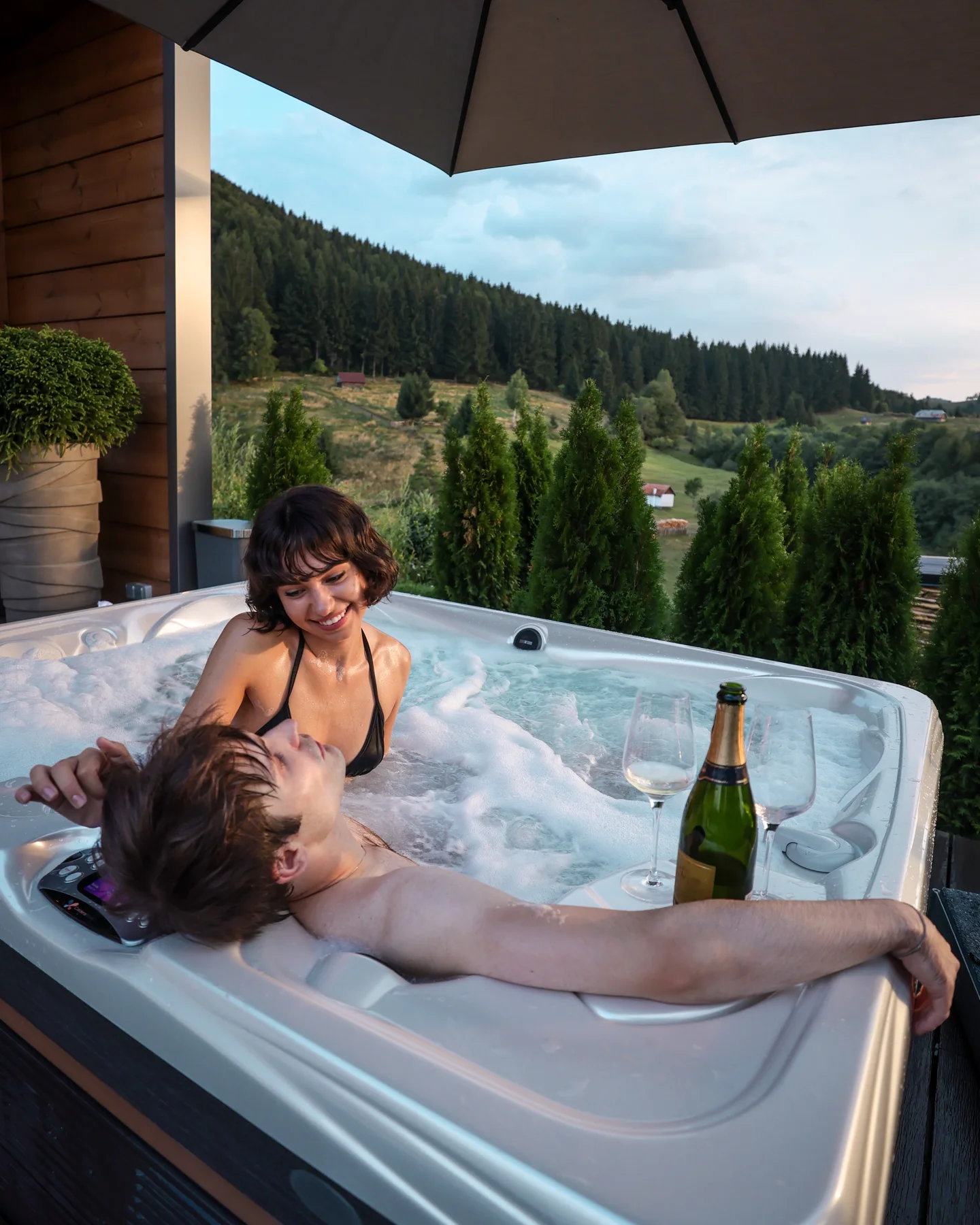 Cuplu într-un jacuzzi pe terasa acoperită Eden Hill, cu o sticlă pe margine și pădure în fundal.